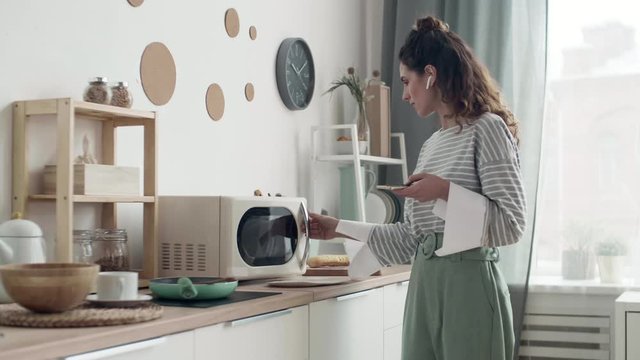 Lockdown of young pretty woman standing in the kitchen, microwaving food while having phone call. Then she taking plate and going away