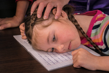 Sad little girl got tired of writing and put her head on the notebook. Mother's hand on her...