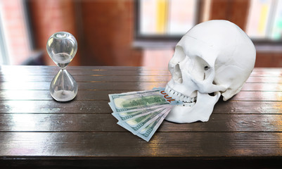 The white skull is lying on the table, holding money in its teeth. Hundred-dollar bills, an hourglass, and a skull on a brown background. A conceptual symbol of bankruptcy, debt repayment, and ruin.