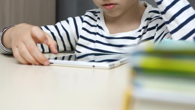 The child is sitting at the table and reading a book using a tablet. The boy is playing, using the app for children. The focus goes from the books on the table to the child's face.
