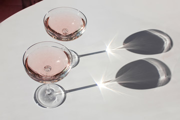 Two glasses of sparkling wine on the pink table in natural sunlight with shadows.