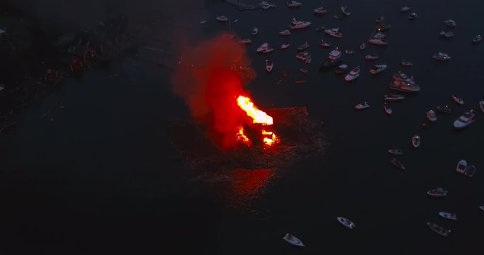 Cinematic, circling, top down aerial drone footage of small boats gathering and witnessing the biggest bonfire event on Earth at Alesund, Norway at dusk. We fly out from the smoke at the beginning.