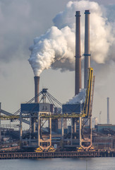 Heavy industry in the port of Ijmuiden Netherlands billowing pollution into a blue sky. A coatal ship passes in the foreground