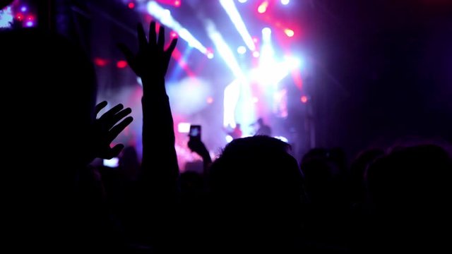 Happy people are watching an amazing musical concert. Merry fans jump and raise their hands up. Crowd of excited fans applauding to popular band performing favorite song. 