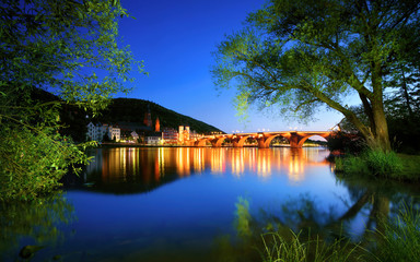 Fototapeta premium Neckar river in Heidelberg, Germany, at dusk, with deep blue sky reflected in the clear water and green trees framing the illuminated Old Bridge