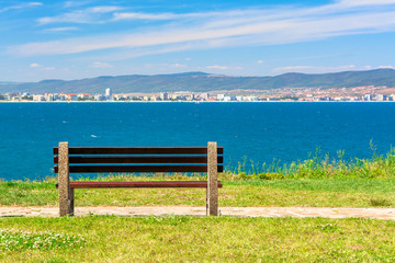 Obraz premium bench on the sunny beach shore. beautiful skyline view from empty park with paved footpath on the seaside. city and mountain in the distance beneath a blue sky with clouds