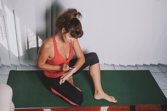 Athletic Pretty Middle Aged Woman In Tight Sportswear, Sitting On Yoga Mat After Workouts, Checking Body Health Indicators On Smart Watch, Fitness Tracker. Sport Activity Indoor