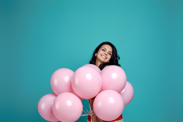 Fototapeta na wymiar Smiling carefree curly girl in dress holding pastel pink air balloons isolated on blue background. Beautiful happy young woman on a birthday party. happiness. space for text