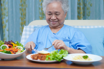 Asian senior or elderly old lady woman patient eating breakfast vegetable healthy food with hope and happy while sitting and hungry on bed in hospital.