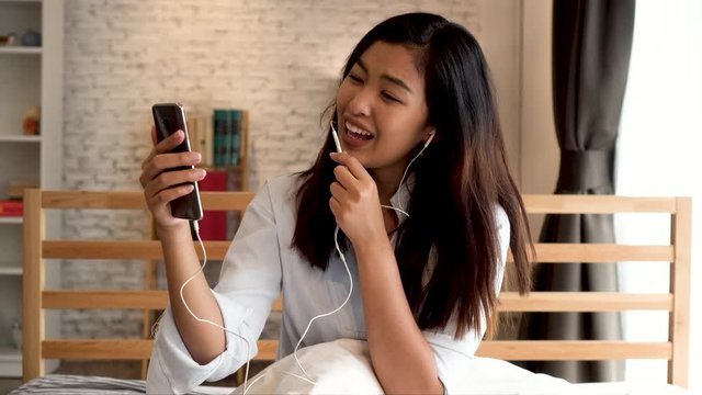 Portrait of happy young Asian girl in casual clothing lying down on bed while making a video call with smartphone in bedroom at home. Video conferencing technology concept.