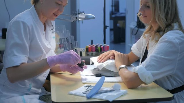 Woman client choosing color of shellac to cover nails in beauty salon, side view. Making manicure for young blonde girl. Beauty industry concept.
