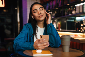 Photo of asian woman using mobile phone and wireless earphones in cafe