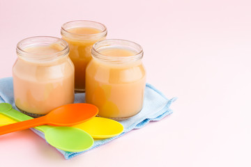 Glass jars of baby food with plastic spoons and blue napkin on pink background