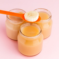 Closeup of glass jars of baby food with banana slice on plastic spoon on pink background