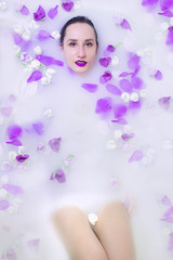 Obraz na płótnie Canvas Young beautiful woman with bright violet make-up in milk bath with violet and white flowers. Healthy dewy skin. Fashion model girl, spa and skin care concept. Spring colors. vertical photo