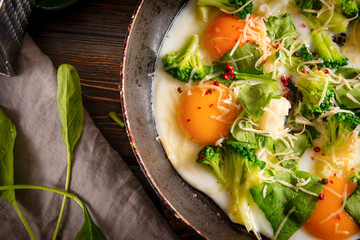 eggs with cheese, broccoli, spinach in a pan on a dark wooden background, protein healthy Breakfast