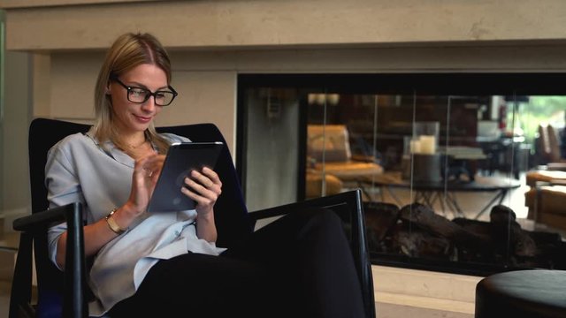 Millennial female in eyewear for vision correction reading received email on modern touch pad, caucasian young smiling woman browsing network on digital tablet while sitting on cozy chair in interior
