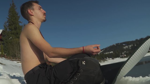 Young man with no shirt breathing heavily, sitting on yoga mat in snowy landscape beside frozen mountain lake prepares for ice bath. Man hyperventilating in winter mountain scene. 