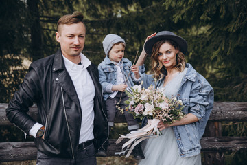 Stylish family in the mountains. A guy in a leather jacket, a girl in a wedding dress and a hat with his son stand under a fir tree near a wooden fence and hold a wedding bouquet