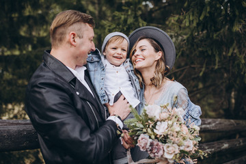 Stylish family in the mountains. A guy in a leather jacket, a girl in a wedding dress and a hat with his son stand under a fir tree near a wooden fence and hold a wedding bouquet