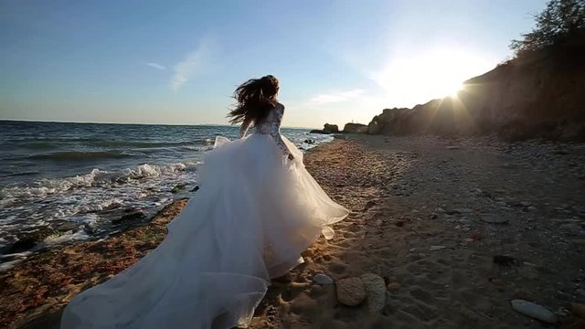 Attractive girl with long hair alone in a white wedding dress walks along the seashore. The bride runs at sunset. on blue sea background. Slow motion of woman, sexy woman, luxurious, smiling happy on 