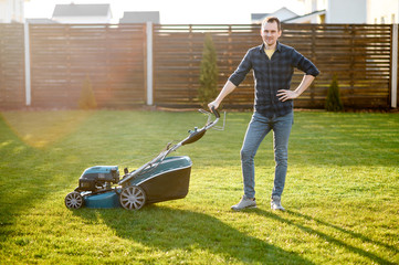 Portrait of guy with a lawn mower on a back yard of private house, he stands and smiles. Care about...