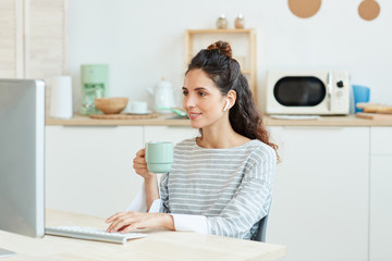 Horizontal medium portrait of young woman using her desktop computer and drinking morning coffee at home