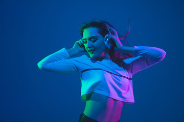 Listening to music. Caucasian young woman's portrait isolated on blue studio background in neon light. Beautiful female model. Concept of human emotions, facial expression, sales, ad, youth culture.