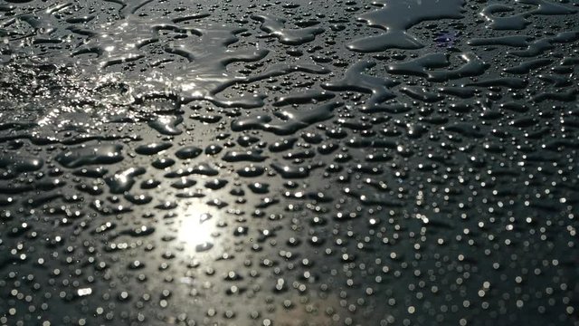 Light rain dripping on a shiny smooth floor with small formed puddles, close view. Reflection of the sun in drops of water. Sunny and rainy weather. Water pours onto the surface of graphite color.