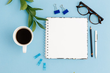 Blank white notebook, leaves, pencil, pen, diary, cup of coffee, paper clips, glasses on colorful blue table. Stylish minimalistic workplace concept. Top view with copy space.