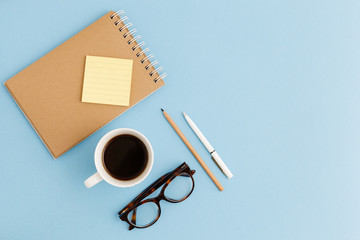 Blank brown vintage notebook, pencil, pen, diary, cup of coffee, paper clips, glasses on colorful blue table. Stylish minimalistic workplace concept. Top view with copy space. 