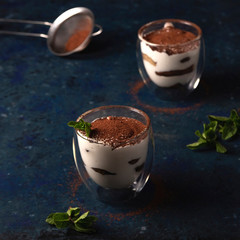 Tiramisu in a transparent glass with a double bottom with cocoa and mint on a blue dark background.