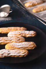 Homemade Savoyardi cookies on a black plate next to a baking sheet and icing sugar.
