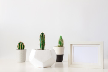 Desk on a white empty wall background with three cactuses in a geometric pot and a frame mockup. Copy space.