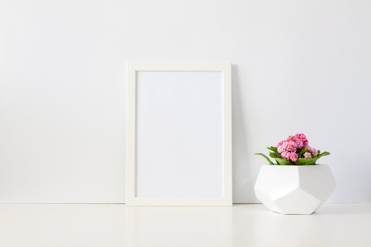 Desk against a white empty wall with a plant with pink flowers in a geometric pot and a frame mockup. Copy space.