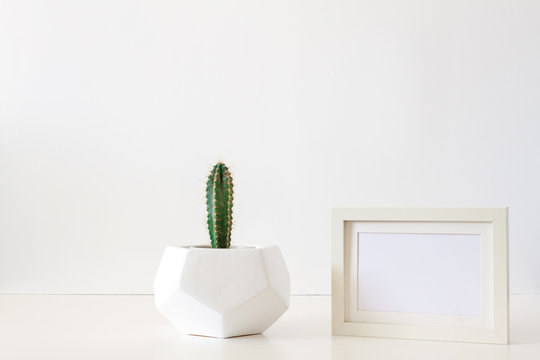 A desk against a white empty wall with a single cactus in a geometric pot and a frame mockup. Copy space.