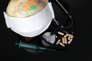 Coronavirus pandemic in world, global quarantine during the COVID-19. Medical mask on globe, stethoscope, syringe and pills on black glass table