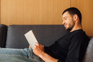 side view of handsome and bearded man reading book in living room