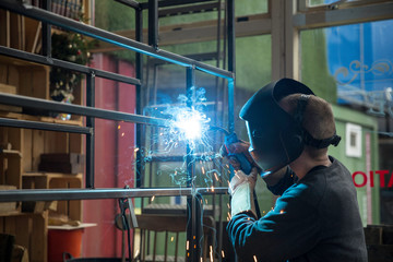 Fototapeta na wymiar Focused welder designer wearing a helmet and working on a project by welding a metal construction while making welding sparks and smoke in his workshop horizontal