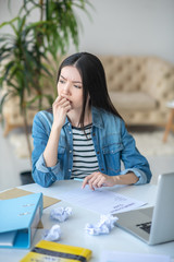 Sad dark-haired female sitting at her desk with unemployment claim, closing her mouth with her hand