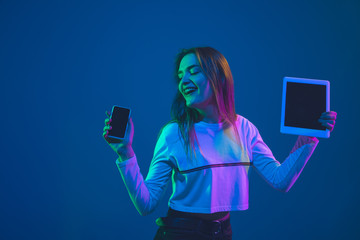 Showing devices. Caucasian young woman's portrait isolated on blue studio background in neon light. Beautiful female model. Concept of human emotions, facial expression, sales, ad, youth culture.