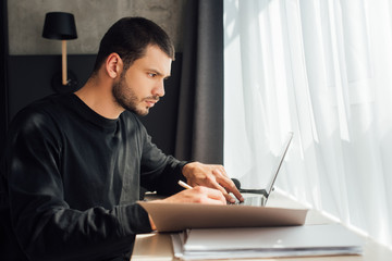 selective focus of bearded freelancer using laptop and holding pencil near folder at home