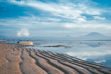 Sanur sandy beach at dawn. Low tide on the beach. Boat at the shore against the background of mountains and a volcano. Calm sea and reflection in the water.