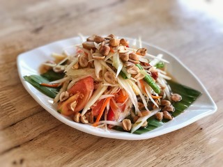 Papaya salad on banana leaf in white plate and put on the wooden table, spicy and sour tasty Thai style food. Thailand famous local food, Asian restaurant concept. Selective focus blur background.