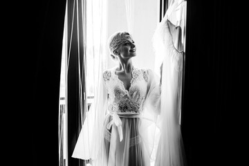 Beautiful bride standing by the window and looking at her wedding dress,  black and white photo