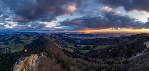 Tourists on a hiking route at the top of the mountain in Jura mountains conteemplating dramatic sunset and colorful rain over the Swiss Central Valley in January with Alps mountains on the horizon