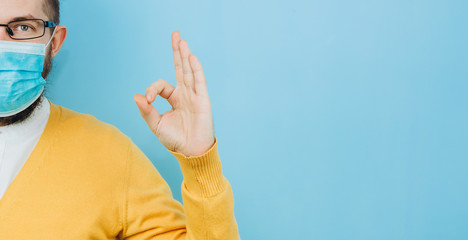 A young man in a medical mask on a blue background. A man in a yellow cardigan shows a hand gesture OK. We're going to beat coronavirus. The COVID-2019 epidemic. Positive boy.