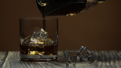 Pouring alcohol drink whiskey, cognac into glass with ice. Dark background.