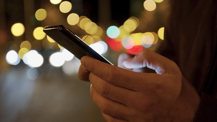 Closeup of male hand typing a cell phone screen and web surfing while standing near a night road.