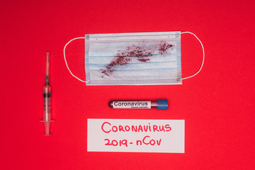 Top view of test tube with blood sample near medical mask with blood, syringe and card with coronavirus 2019-nCov lettering on red surface